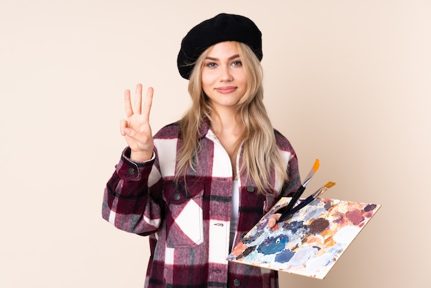 Teenager-Künstlermädchen, das eine Palette auf der blauen Wand glücklich hält und drei mit den Fingern zählt