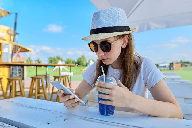 Teenager, Lifestyle, Sommer, Urlaub. Teenager-Mädchen, das einen Videoanruf auf einem Smartphone macht, das an einem Tisch in einem Sommercafé im Freien mit Beerengetränk sitzt