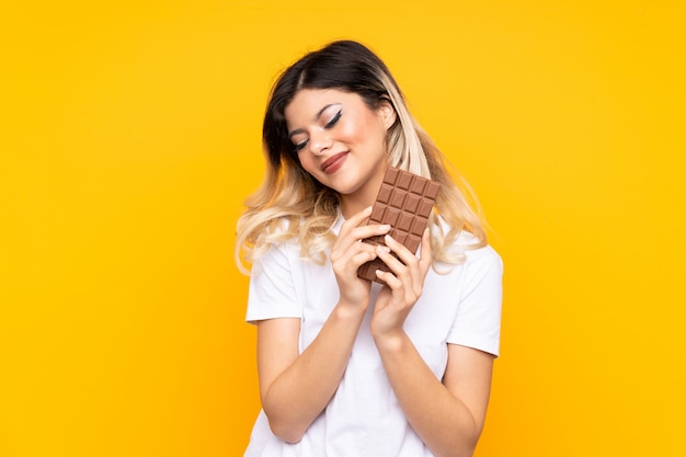 Teenager-Mädchen auf gelber Wand, die eine Schokoladentafel nimmt und glücklich