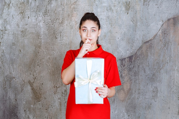 Teenager-Mädchen, das eine blaue Geschenkbox hält, die mit weißem Band umwickelt ist und gestresst oder verängstigt aussieht.