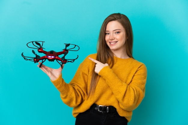 Teenager-Mädchen, das eine Drohne über isoliertem blauem Hintergrund hält und auf die Seite zeigt, um ein Produkt zu präsentieren