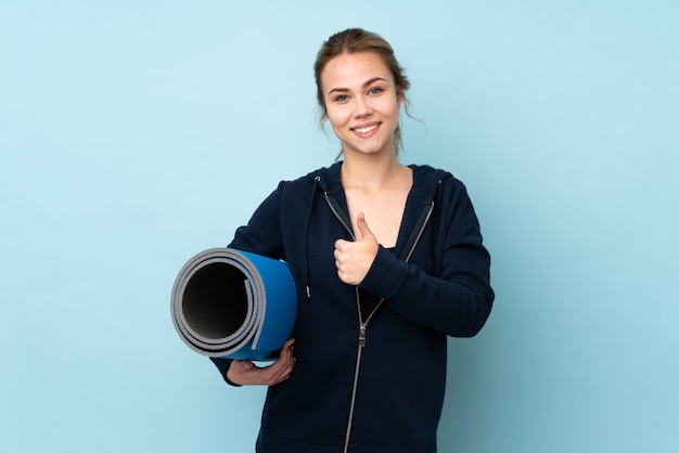 Teenager-Mädchen, das Matte lokalisiert auf Blau hält und eine Daumen hoch Geste gibt