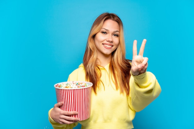 Teenager-Mädchen, das Popcorn über lokalisiertem blauem Hintergrund hält, der lächelt und Siegeszeichen zeigt