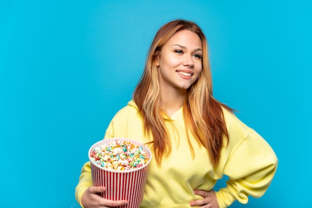 Teenager-Mädchen, das Popcorn über lokalisiertem blauem Hintergrund hält, der mit Armen an der Hüfte aufwirft und lächelt