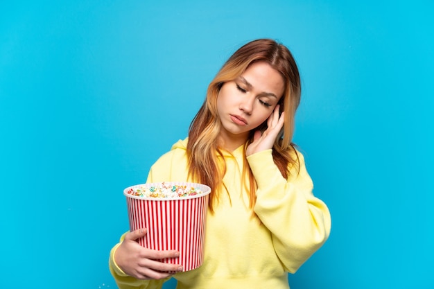 Teenager-Mädchen, das Popcorn über lokalisiertem blauem Hintergrund mit Kopfschmerzen hält