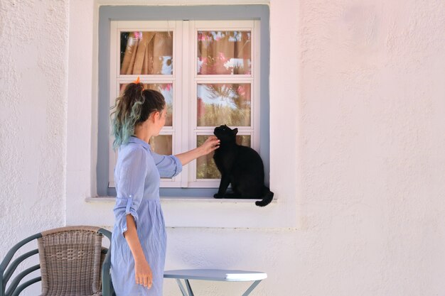 Teenager-Mädchen, das schöne schwarze Katze berührt, die draußen auf der Fensterbank sitzt