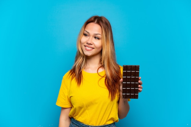 Teenager-Mädchen, das Schokolade über isoliertem blauem Hintergrund hält und zur Seite schaut und lächelt