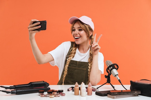 Teenager-Mädchen, das Selfie auf dem Smartphone macht und eine Blogsendung über neue Kosmetikprodukte aufnimmt, die auf orangefarbener Wand isoliert sind?