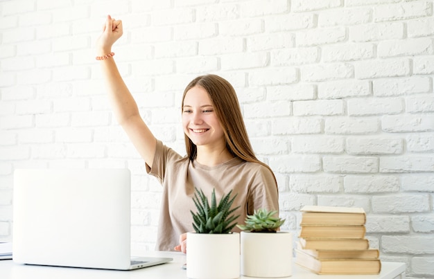 Teenager-Mädchen, das von zu Hause aus am Schreibtisch mit erhobenem Arm sitzt