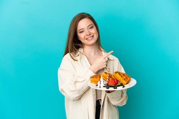 Teenager-Mädchen, das Waffeln über isoliertem blauem Hintergrund hält und auf die Seite zeigt, um ein Produkt zu präsentieren