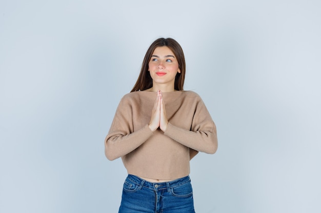 Teenager-Mädchen drückt die Hände zusammen, während sie in Pullover, Jeans betet und hoffnungsvoll aussieht. Vorderansicht.