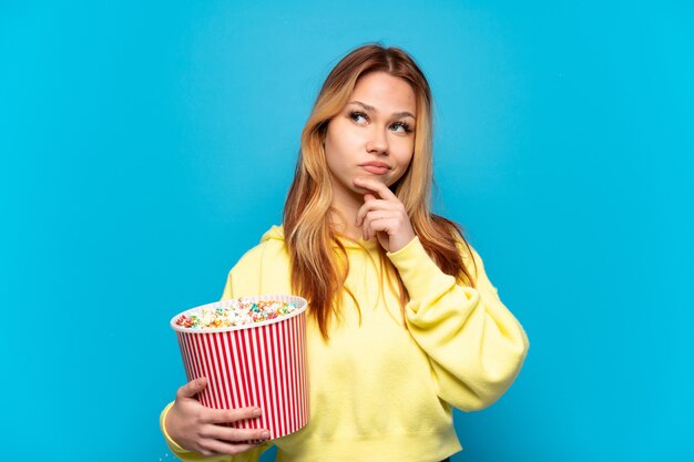 Teenager-Mädchen hält Popcorn über isoliertem blauem Hintergrund und schaut nach oben