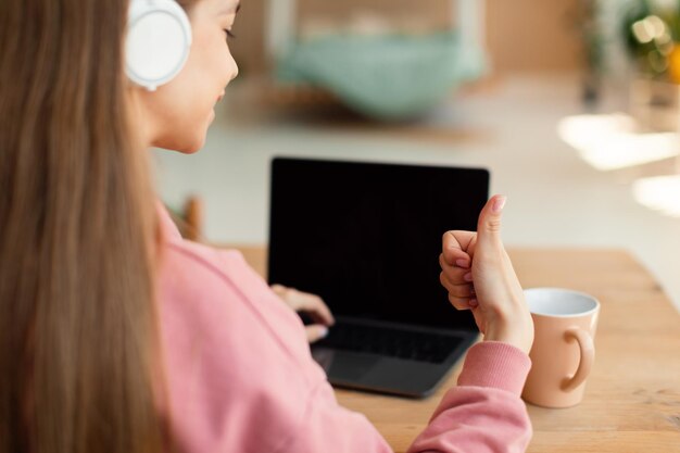 Teenager-Mädchen in drahtlosen Kopfhörern, die mit Laptop mit leerem Bildschirm lernen und den Daumen nach oben zeigen, während sie zu Hause am Schreibtisch sitzen
