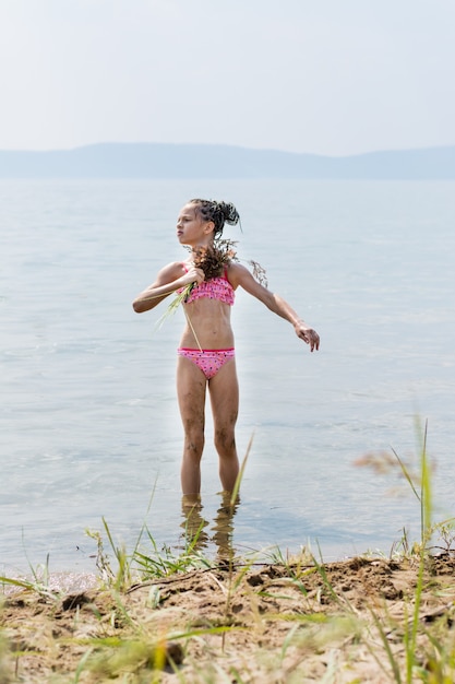 Teenager-Mädchen in einem Badeanzug, der sich im Sommer mit einem Grasbesen fächert, der Badeverfahren am sonnigen Flussufer imitiert?