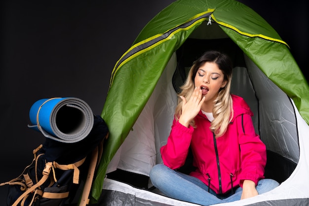 Teenager-Mädchen in einem grünen Zelt des Campings lokalisiert auf schwarzem Hintergrund, der gähnt und weit geöffneten Mund mit Hand bedeckt