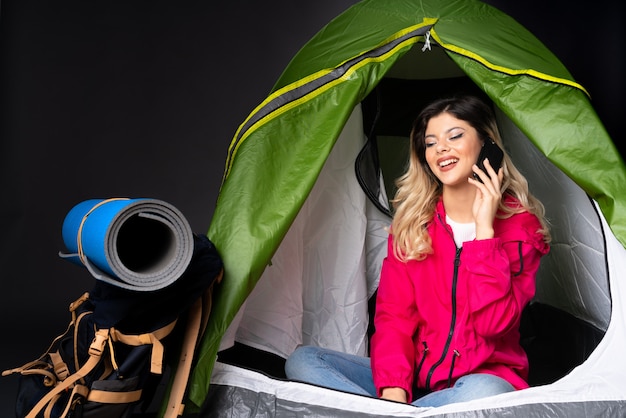 Teenager-Mädchen in einem grünen Zelt des Campings lokalisiert auf schwarzer Wand, die ein Gespräch mit dem Mobiltelefon hält