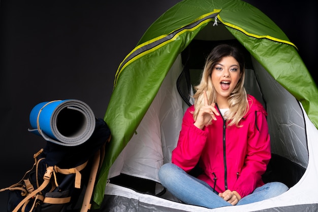 Teenager-Mädchen in einem grünen Zelt des Campings lokalisiert auf schwarzer Wand überrascht und nach vorne zeigend