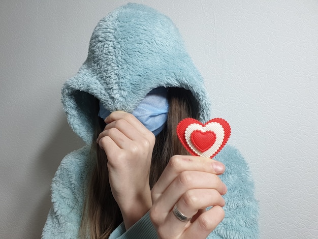 Teenager-Mädchen in einer medizinischen Maske hält ein rotes Herz in der Hand, Valentinstag in Quarantäne