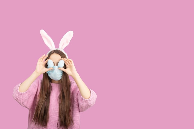 Teenager-Mädchen in Hasenohren mit Ostereiern in einer medizinischen Maske auf einem rosafarbenen Hintergrundkonzept des Gesundheitsschutzes vor covid19 während Ostern