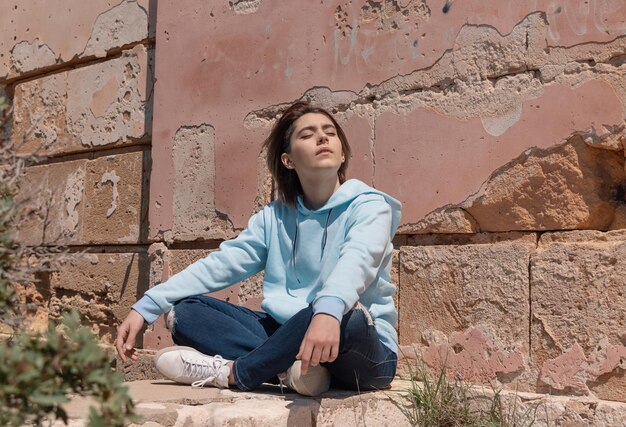 Teenager-Mädchen in hellblauem Hoodie, das auf ihren Hüften in der Nähe einer Wand sitzt