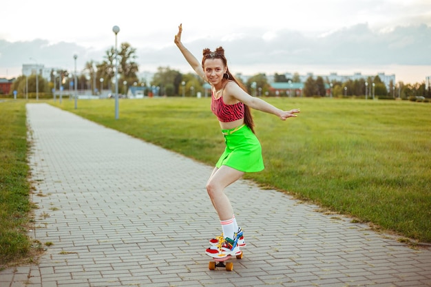 Teenager-Mädchen in heller, stilvoller Kleidung, die Skateboard entlang der Straße im Stadtpark fährt