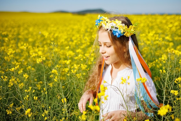 Teenager-Mädchen in Kleid und ukrainischem Kranz mit Linsen auf dem Kopf im Rapsfeld unter einem strahlend blauen Himmel