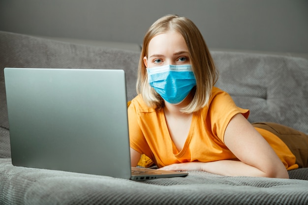 Teenager-Mädchen in Maske machen Online-Lernausbildung über Laptop-Kovid-19-Sperrzeit. Remote-Arbeit in der Coronavirus-Pandemie. Frau in medizinischer Schutzmaskenarbeit mit Laptop im Home Office liegt auf der Couch.