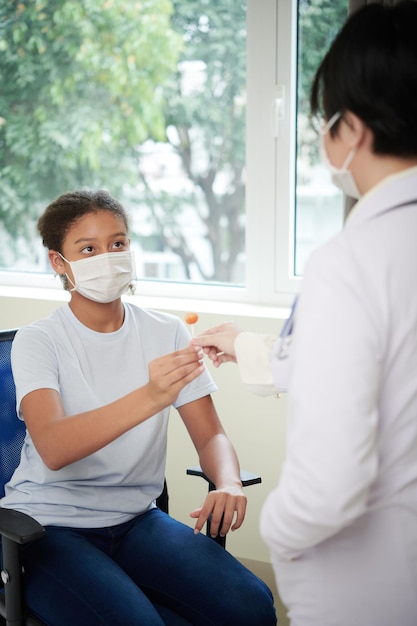 Teenager-Mädchen in Maske sitzt auf einem Stuhl und bekommt die Süßigkeiten von der Ärztin während der ärztlichen Untersuchung im Krankenhaus