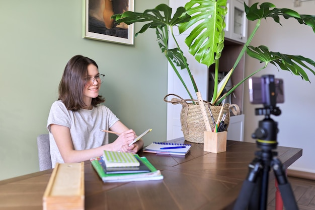 Teenager-Mädchen lernt zu Hause, sitzt an einem Tisch mit Büchern und Notizbüchern mit einem Smartphone, Videoanruf-Konferenz-Fernunterricht. Technologien in der Bildung, Online-Lernen