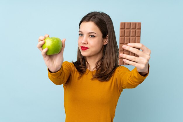 Teenager-Mädchen lokalisiert auf blauer Wand, die eine Schokoladentafel in einer Hand und einen Apfel in der anderen nimmt