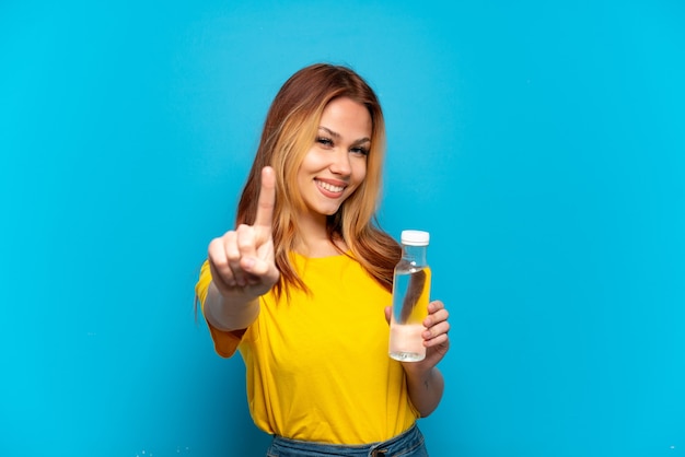 Teenager-Mädchen mit einer Flasche Wasser über isoliertem blauem Hintergrund, der einen Finger zeigt und hebt