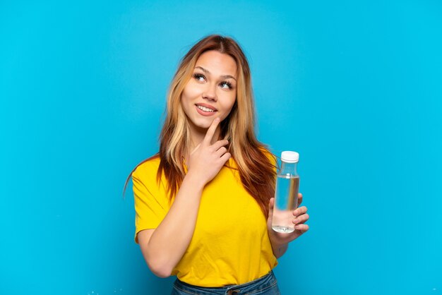 Teenager-Mädchen mit einer Flasche Wasser über isoliertem blauem Hintergrund, der lächelnd nach oben schaut