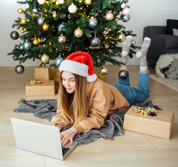 Teenager-Mädchen mit Geschenken und Laptop nahe dem Weihnachtsbaum. Wohnzimmerinnenraum mit Weihnachtsbaum und Dekorationen. Neujahr. Geschenk Geben.