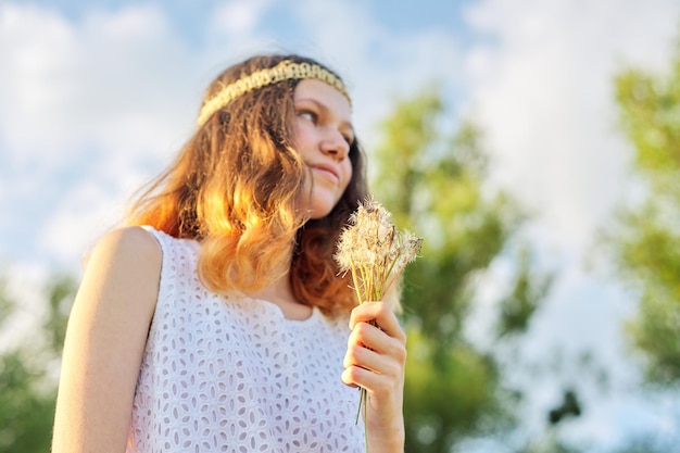 Teenager-Mädchen mit Hippie-Frisur, die trockenen Löwenzahn in der Hand hält. Sommerferien, Urlaub, Natur, Schönheit, Lifestyle. Himmel, Wiese, Hintergrundbeleuchtung bei Sonnenuntergang