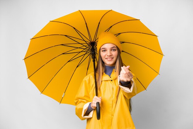 Teenager-Mädchen mit regenfestem Mantel und Regenschirm über isoliertem weißem Hintergrund, Geldgeste