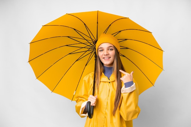 Teenager-Mädchen mit regenfestem Mantel und Regenschirm vor isoliertem weißem Hintergrund mit einer Daumen-hoch-Geste