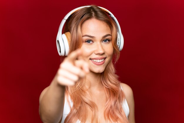 Teenager-Mädchen mit rosa Haaren über isolierter roter Wand, die Musik hört und nach vorne zeigt