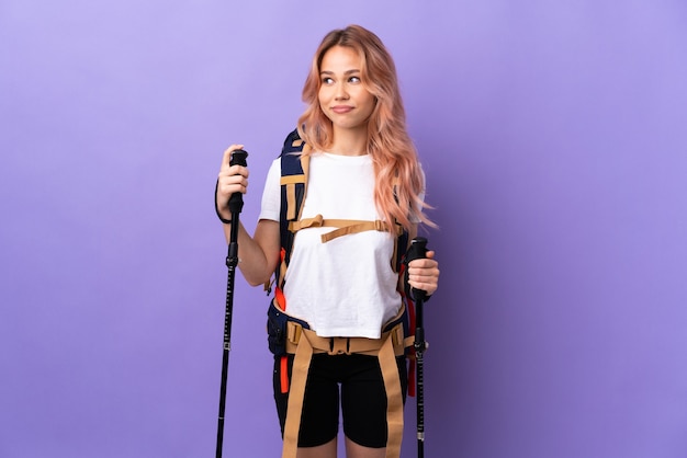Teenager-Mädchen mit Rucksack und Trekkingstöcken über isolierte lila Wand, die Zweifel beim Betrachten der Seite hat