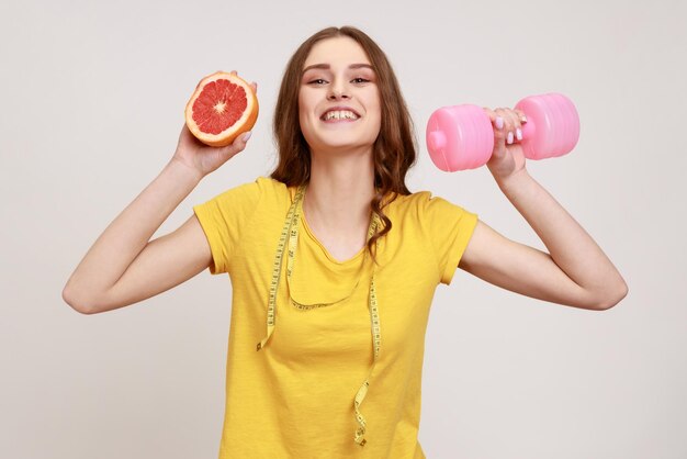 Teenager-Mädchen mit zahnigem gelbem T-Shirt, das rosafarbene Hantel und die Hälfte der Grapefruit hält und sich für Sport und gesunden Lebensstil entscheidet. Studioaufnahme im Innenbereich isoliert auf grauem Hintergrund.