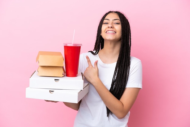 Teenager-Mädchen mit Zöpfen, die Pizza und Burger über isoliertem rosa Hintergrund halten, der auf die Seite zeigt, um ein Produkt zu präsentieren