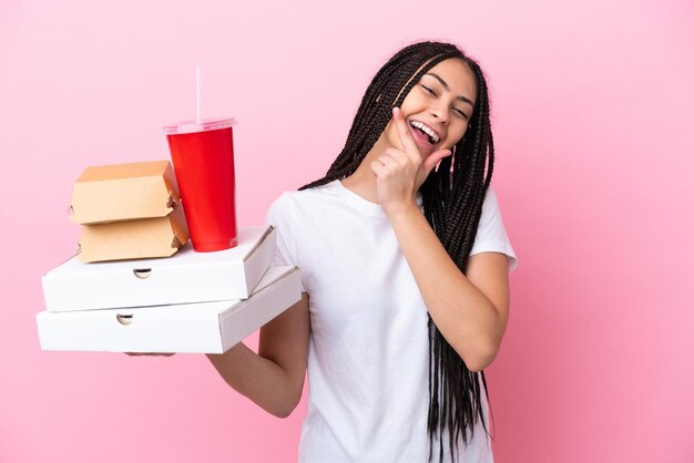 Teenager-Mädchen mit Zöpfen, die Pizza und Burger über isoliertem rosa Hintergrund halten, glücklich und lächelnd