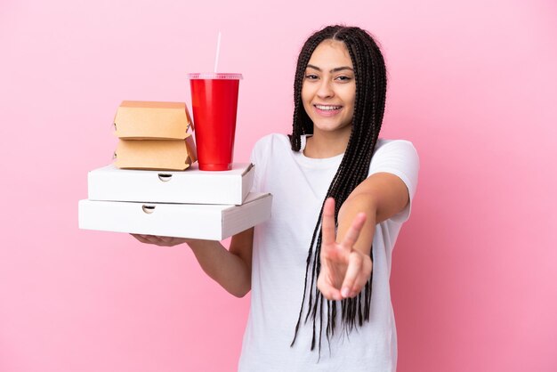 Teenager-Mädchen mit Zöpfen, die Pizza und Burger über isoliertem rosa Hintergrund halten, lächeln und Siegeszeichen zeigen