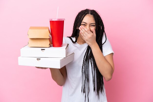 Teenager-Mädchen mit Zöpfen, die Pizzen und Burger über isoliertem rosa Hintergrund halten, glücklich und lächelnd, den Mund mit der Hand bedeckend