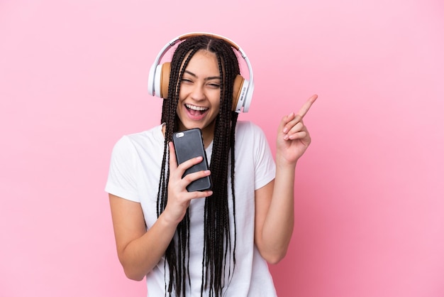 Teenager-Mädchen mit Zöpfen vor isoliertem rosa Hintergrund, das Musik mit einem Handy hört und singt