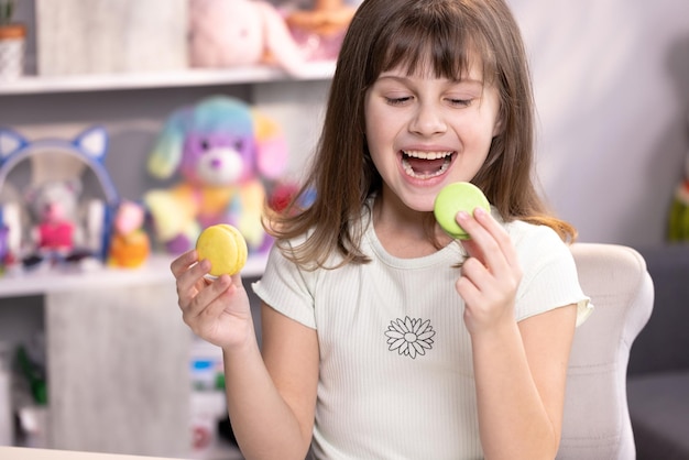 Teenager-Mädchen spielt mit Dessert-Macarons, die die Kekse wie eine Brille um die Augen glücklich lächelnd halten