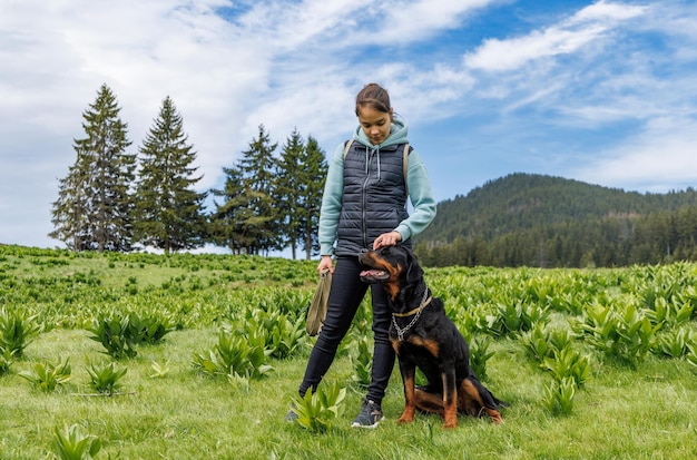 Teenager-Mädchen steht mit Leine in den Händen neben Hund der Rottweiler-Rasse auf der Wiese vor dem Hintergrund von Tannen