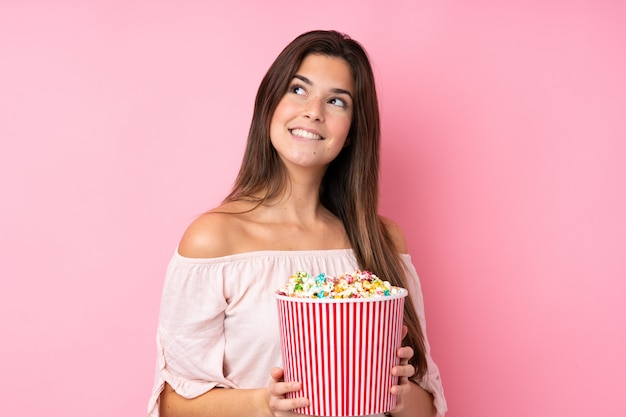 Teenager-mädchen über isolierte rosa wand, die einen großen eimer popcorn hält