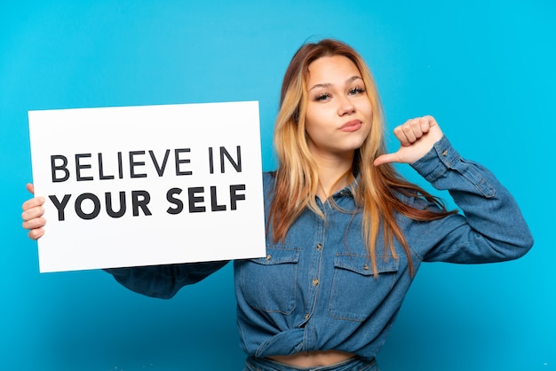 Foto teenager-mädchen über isoliertem blauem hintergrund, das ein plakat mit dem text believe in your self mit stolzer geste hält