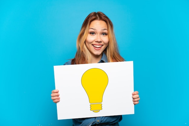 Teenager-Mädchen über isoliertem blauem Hintergrund, der ein Plakat mit Glühbirnensymbol mit glücklichem Ausdruck hält