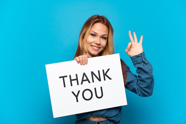 Teenager-Mädchen über isoliertem blauem Hintergrund, der ein Plakat mit Text DANKE mit OK-Zeichen hält